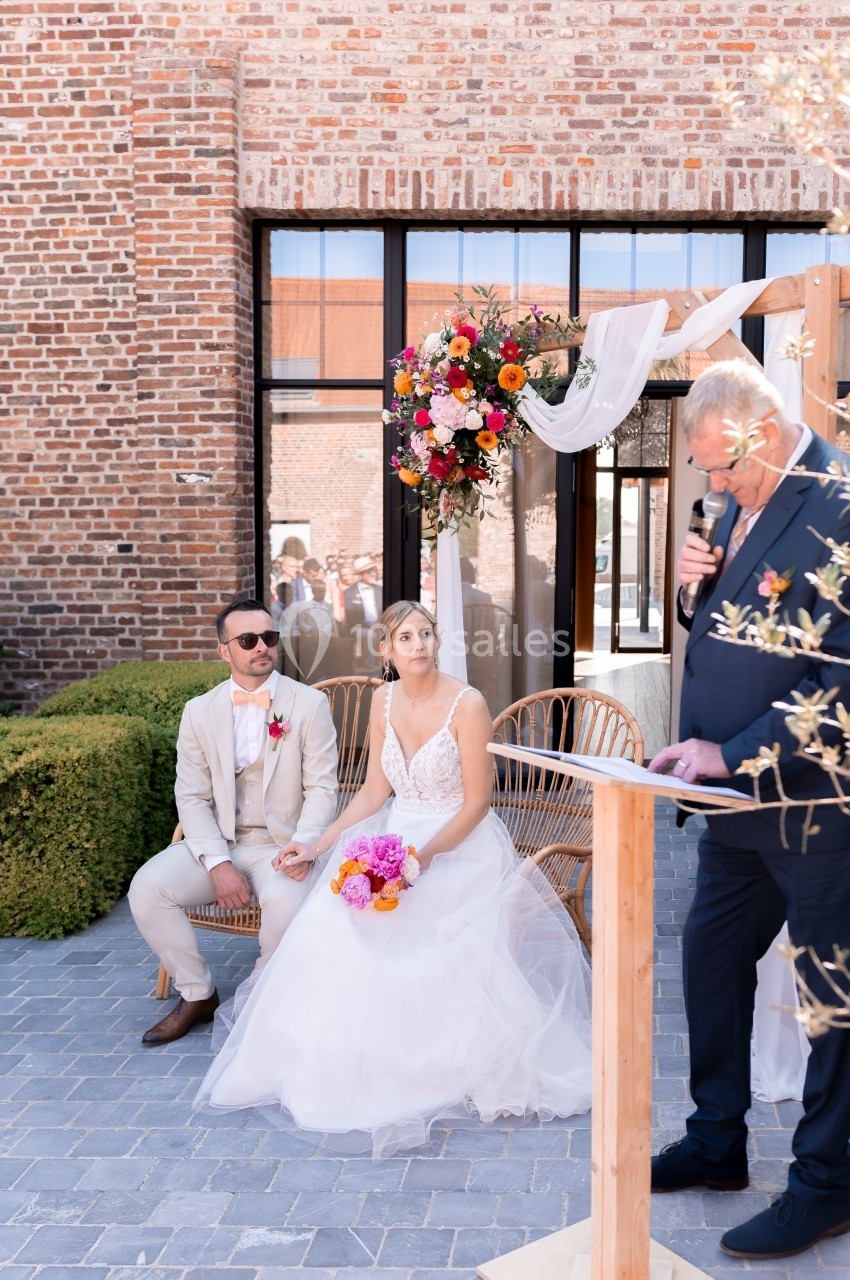 Un couple assis lors d'une cérémonie de mariage en extérieur, écoutant un discours près d'une arche fleurie.