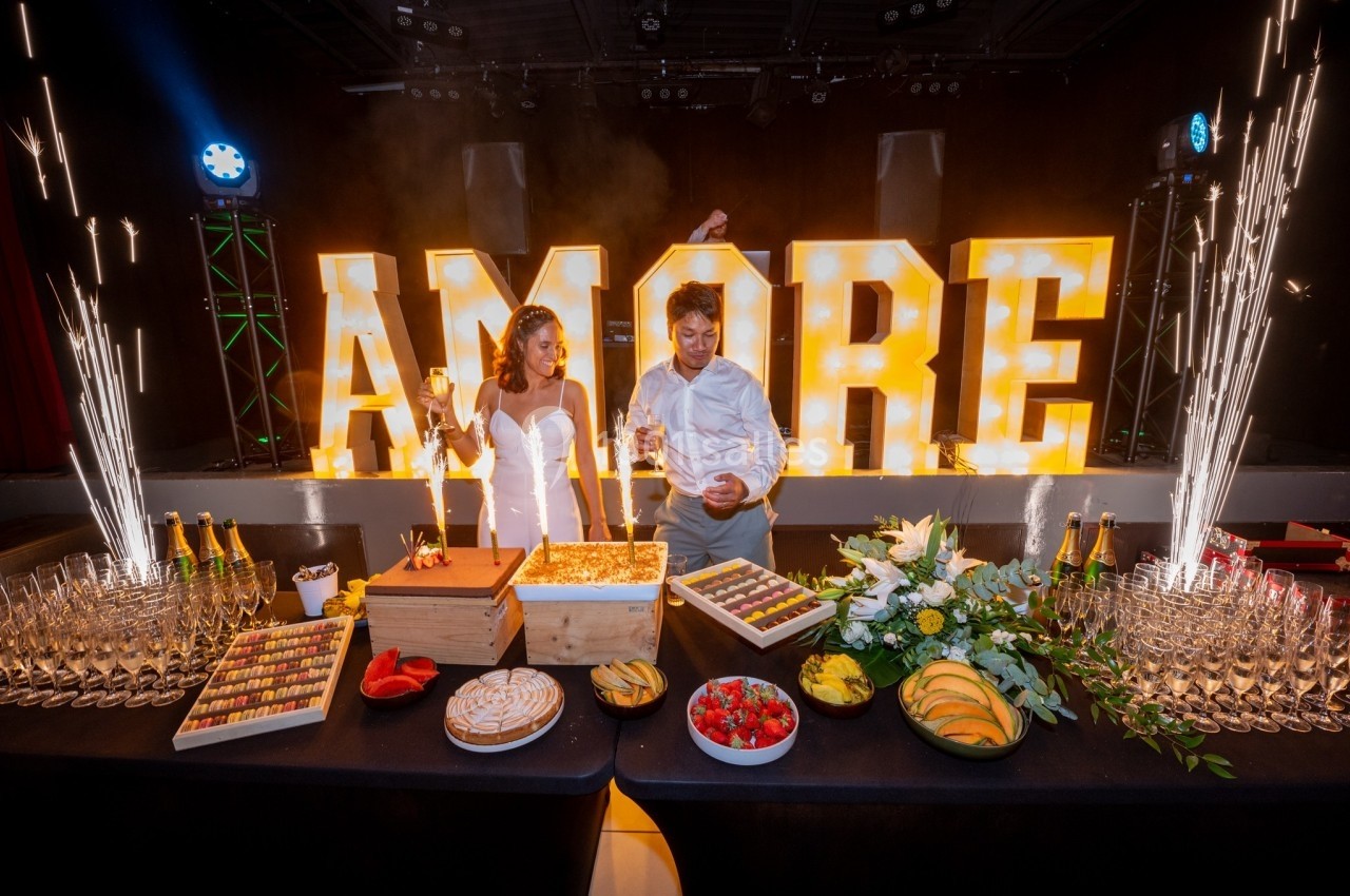 Un couple célèbre son mariage devant une table de desserts et une décoration lumineuse ’AMORE’.