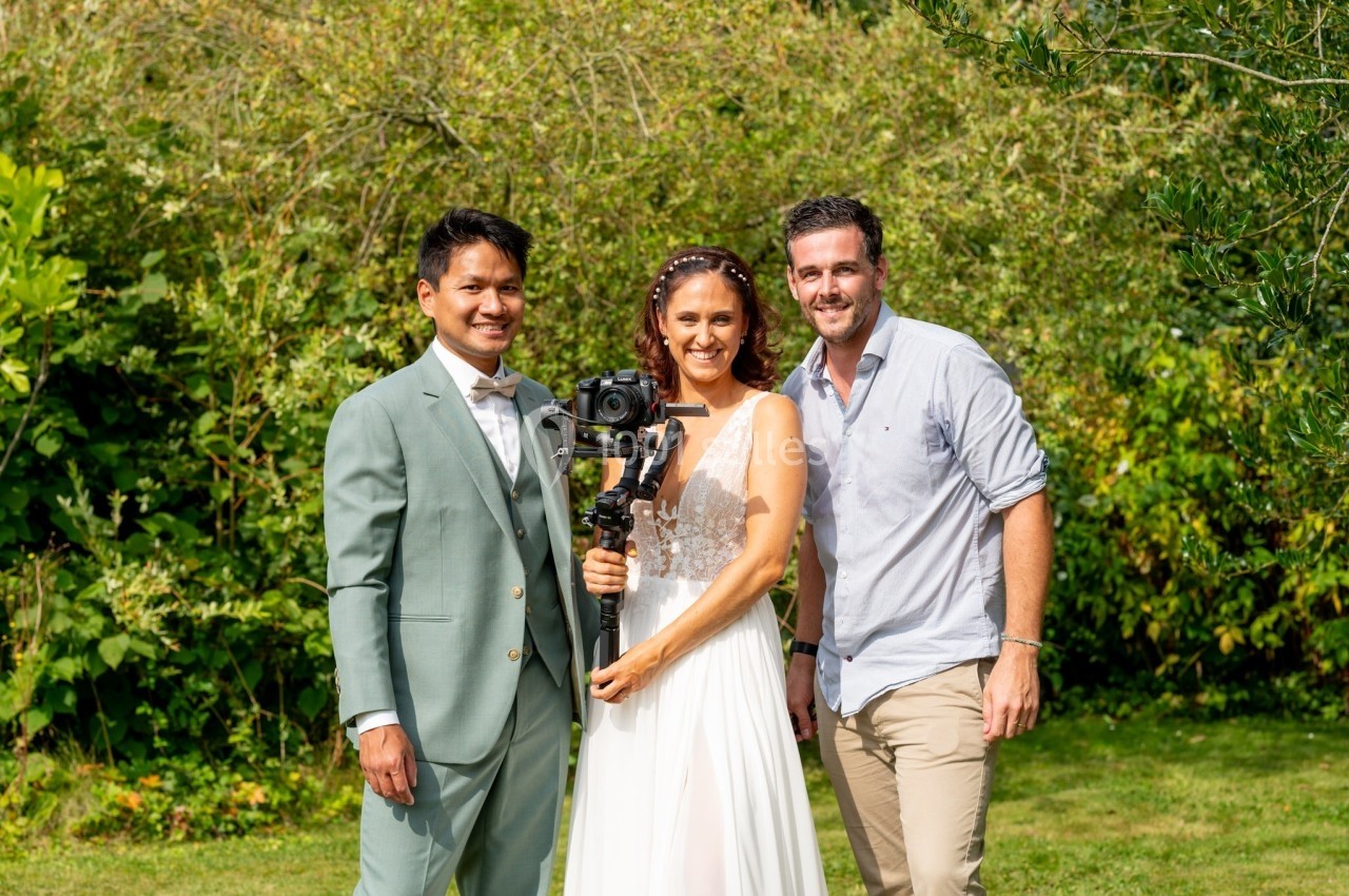Un couple en tenue de mariage pose avec un vidéaste tenant une caméra stabilisée dans un jardin verdoyant.