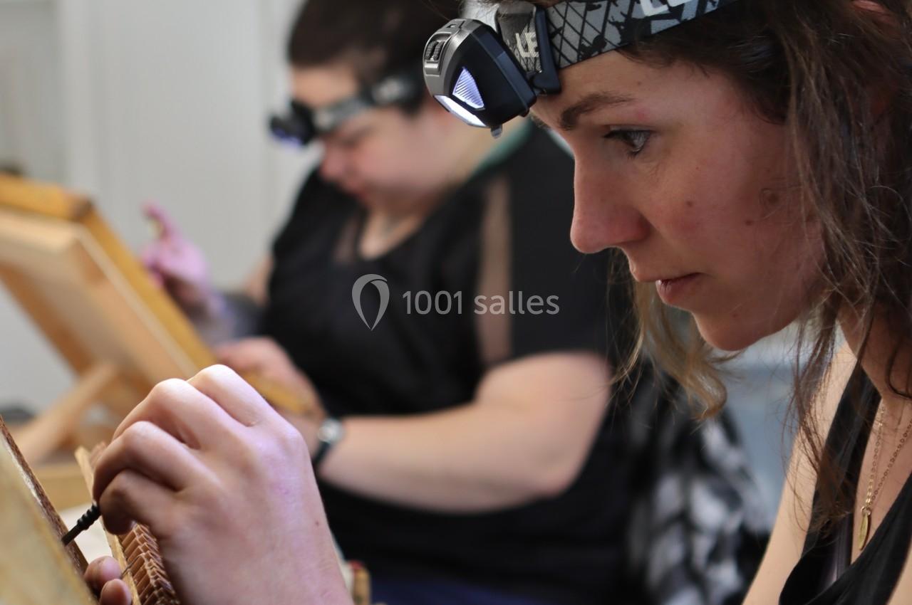 Deux femmes concentrées travaillent sur des cadres en bois, équipées de lampes frontales pour un travail minutieux.