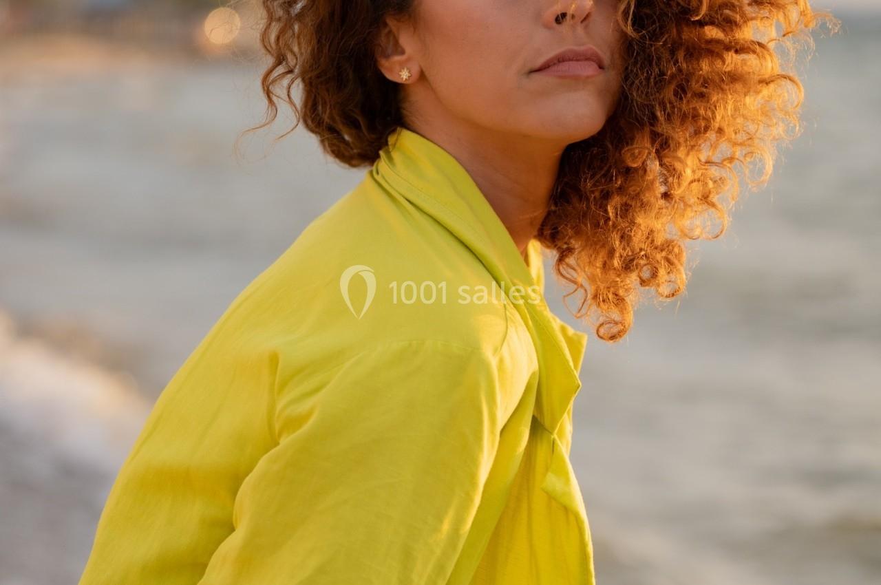 Femme aux cheveux bouclés portant une veste jaune, debout près d'une plage au coucher du soleil.
