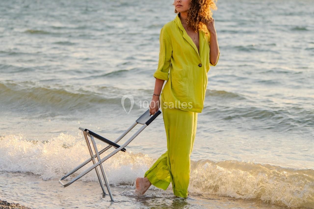 Femme en tenue verte marchant pieds nus sur une plage au bord de l'eau, tenant une chaise pliante.