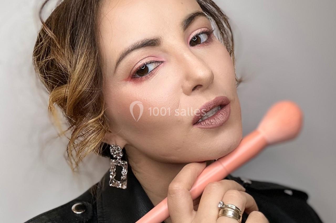 Femme appliquant du maquillage avec un pinceau rose, portant une veste en cuir noir et des boucles d'oreilles.