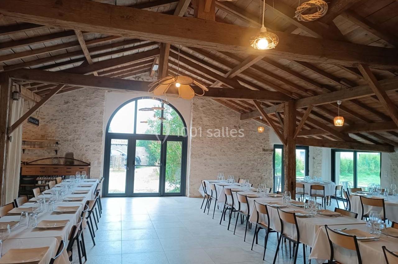 Salle de réception lumineuse avec tables dressées, poutres apparentes et grande baie vitrée donnant sur l'extérieur.