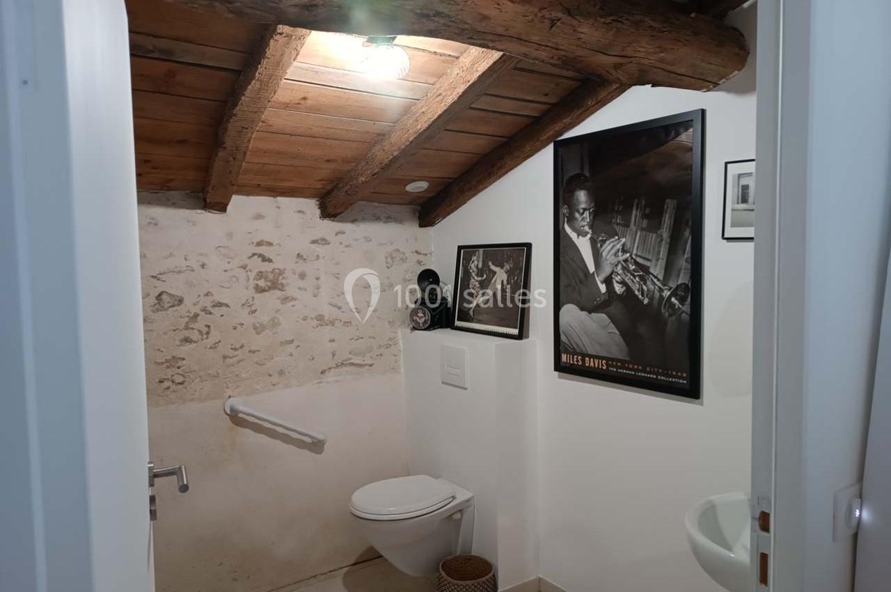 Toilettes sous un plafond en bois incliné, avec murs en pierre, affiches encadrées et un petit lavabo.