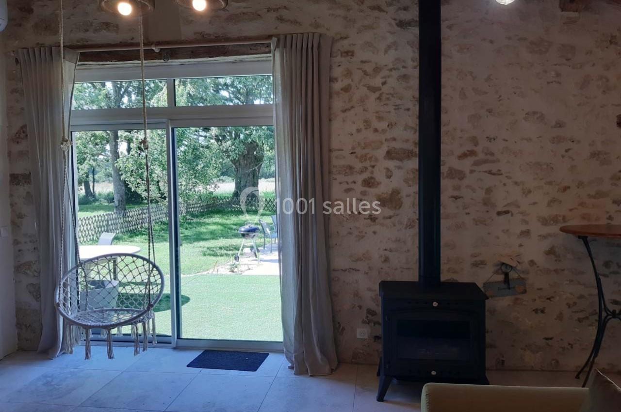 Intérieur lumineux avec mur en pierre, poêle à bois, fauteuil suspendu et baie vitrée donnant sur un jardin verdoyant.