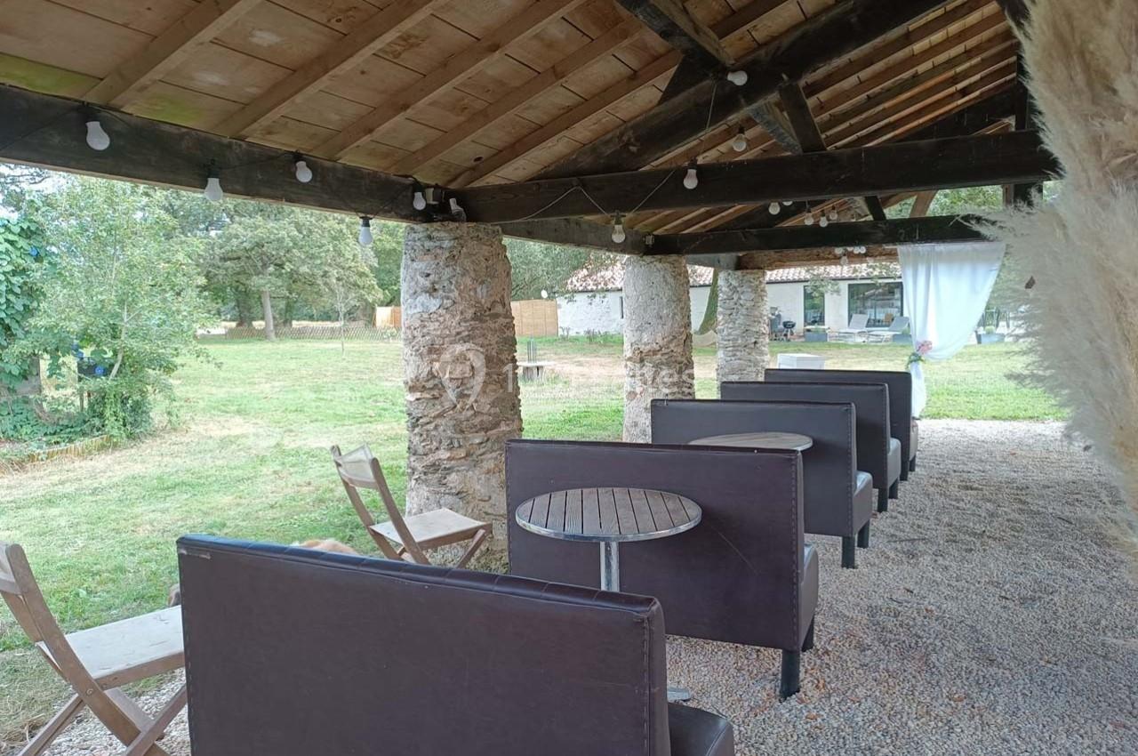 Terrasse couverte avec tables rondes, banquettes marron et chaises en bois, donnant sur un jardin verdoyant.