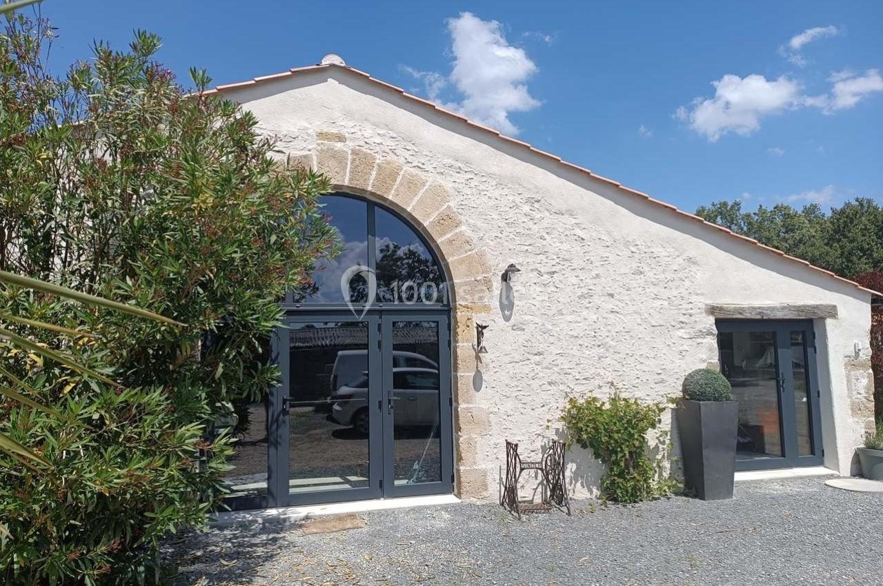 Façade d'une maison en pierre avec grandes baies vitrées, entourée de végétation sous un ciel bleu.