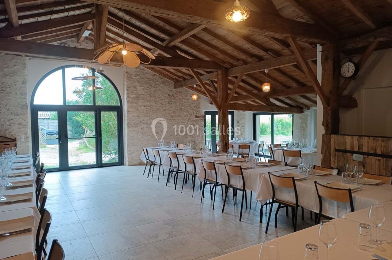 Salle de réception lumineuse avec tables dressées, poutres apparentes et grandes fenêtres donnant sur l'extérieur.