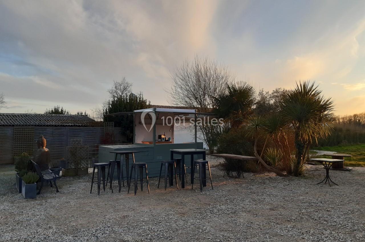 Un bar extérieur en bois avec des tabourets, entouré de végétation et éclairé par une lumière de fin de journée.