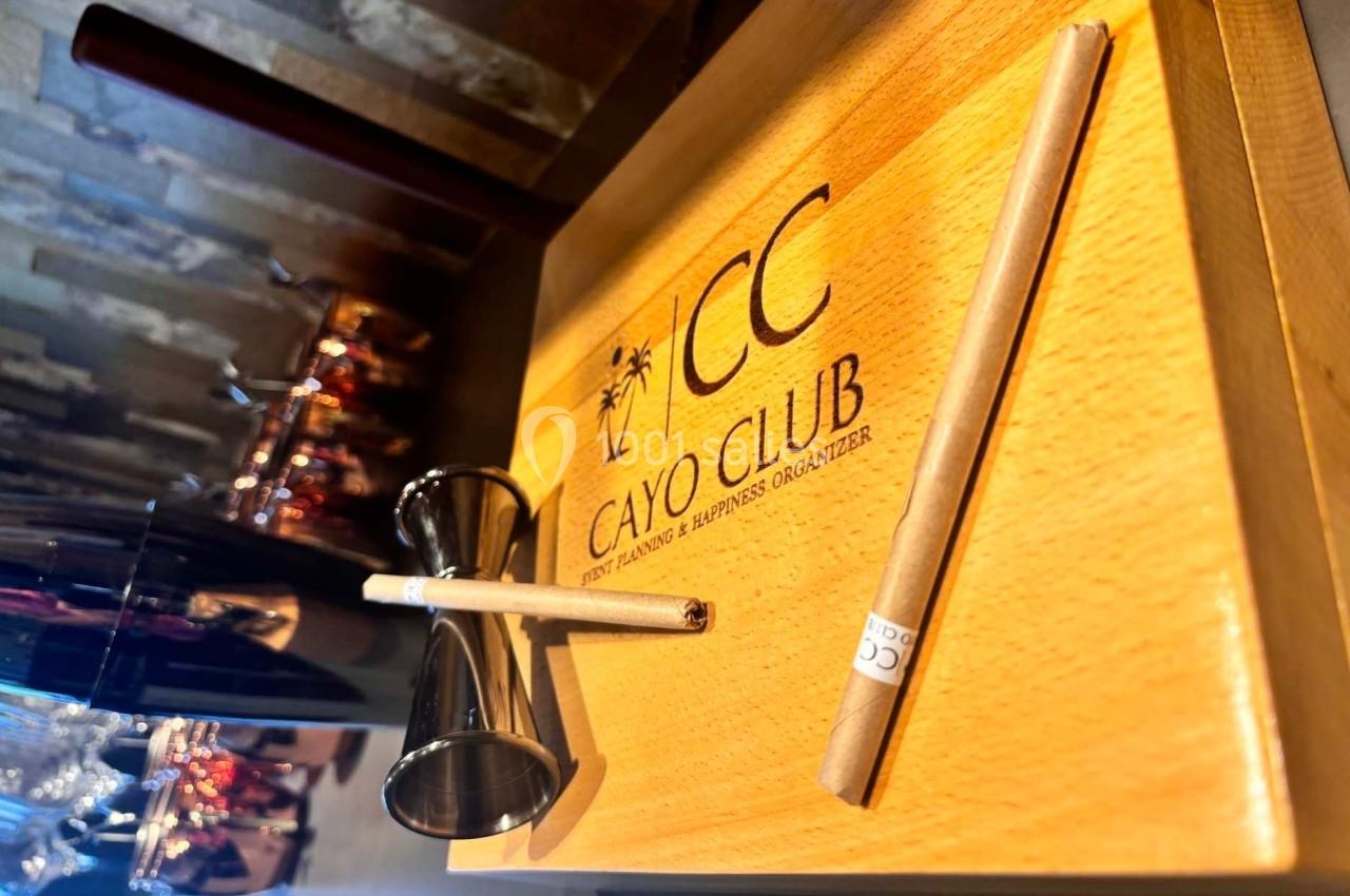 Planche en bois gravée ’Cayo Club’ avec un doseur en métal et deux crayons posés dessus, en arrière-plan des verres.
