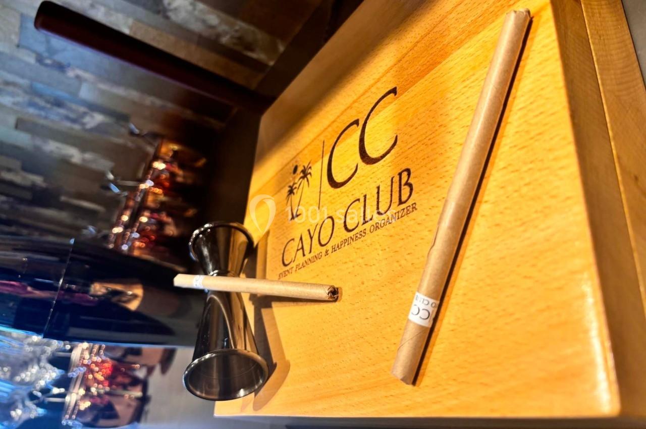 Planche en bois gravée ’Cayo Club’ avec un doseur métallique et un bâtonnet posés dessus, sur un comptoir.