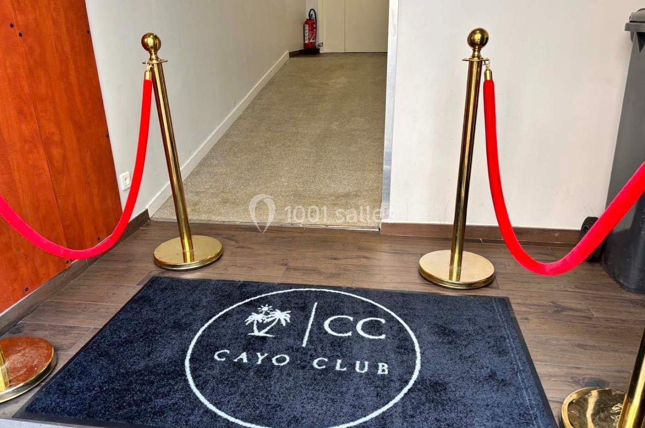 Tapis noir marqué ’Cayo Club’ devant une entrée délimitée par des poteaux dorés et des cordons rouges.