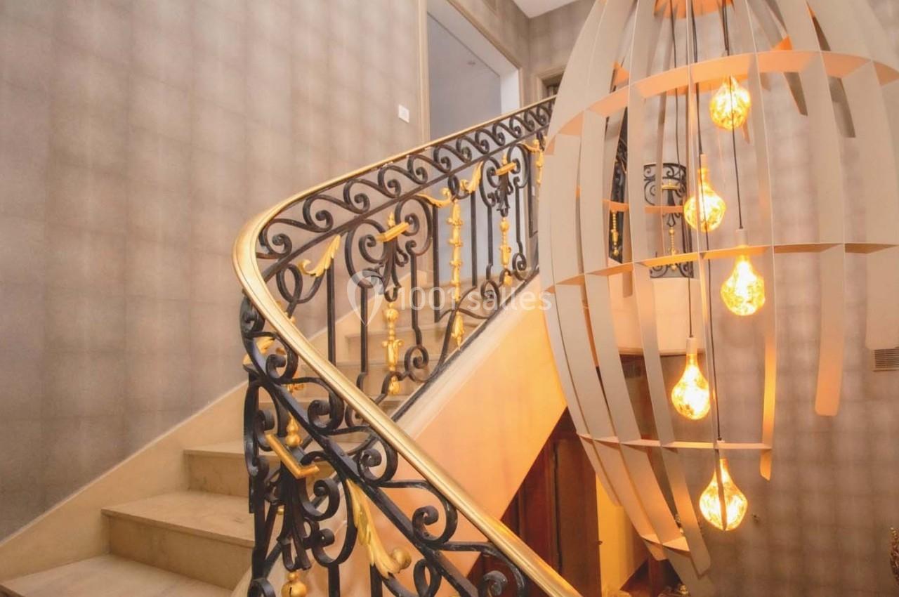 Escalier en marbre avec rampe en fer forgé ornée, éclairé par une suspension moderne avec ampoules apparentes.