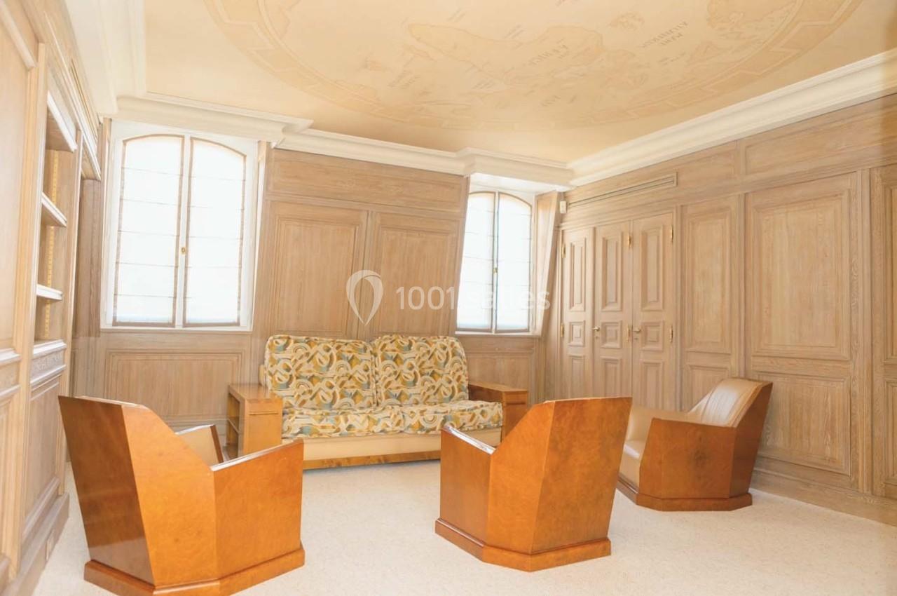 Salle lumineuse avec boiseries murales, plafond décoré, canapé à motifs et fauteuils en bois.
