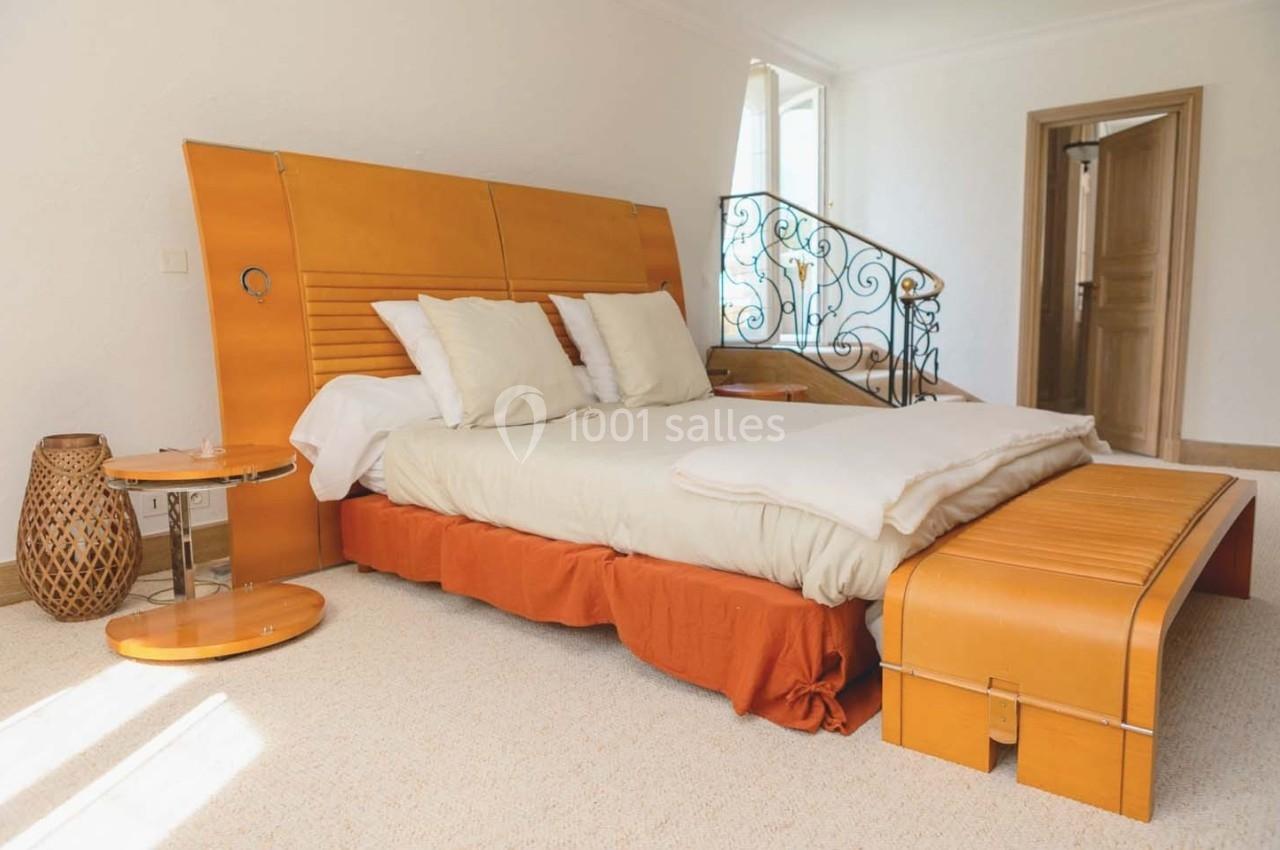 Lit double avec tête de lit en bois, draps clairs et coussins, dans une chambre lumineuse avec escalier en arrière-plan.