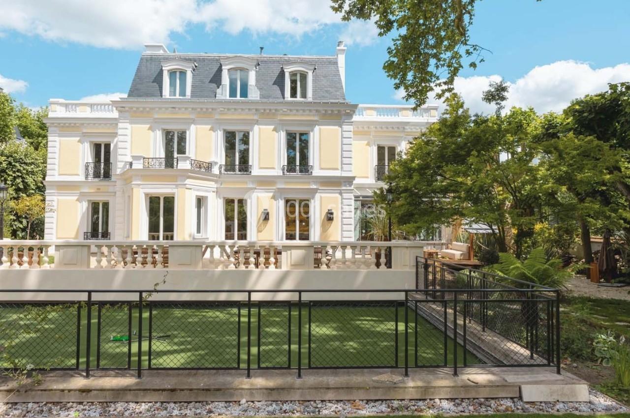 Façade d'une maison bourgeoise à plusieurs étages, entourée d'un jardin avec pelouse et clôture métallique.
