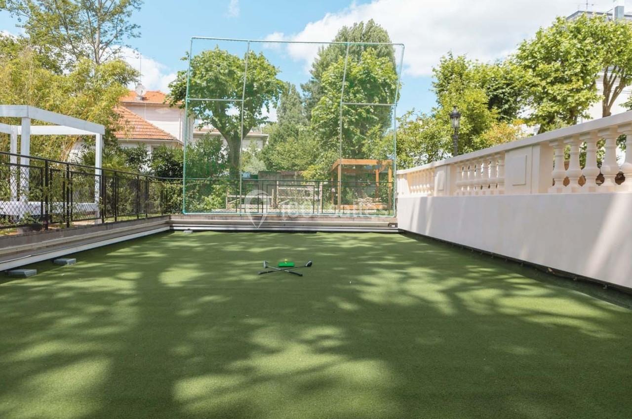 Terrain de pétanque en gazon synthétique entouré de clôtures et d'arbres, sous un ciel dégagé.