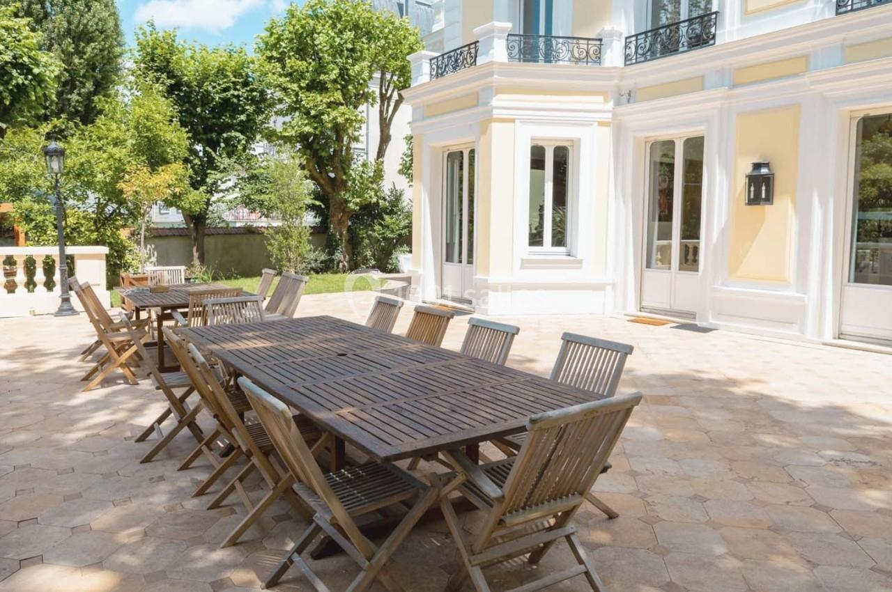 Terrasse pavée avec une grande table en bois et plusieurs chaises, entourée d'arbres et d'une façade claire.