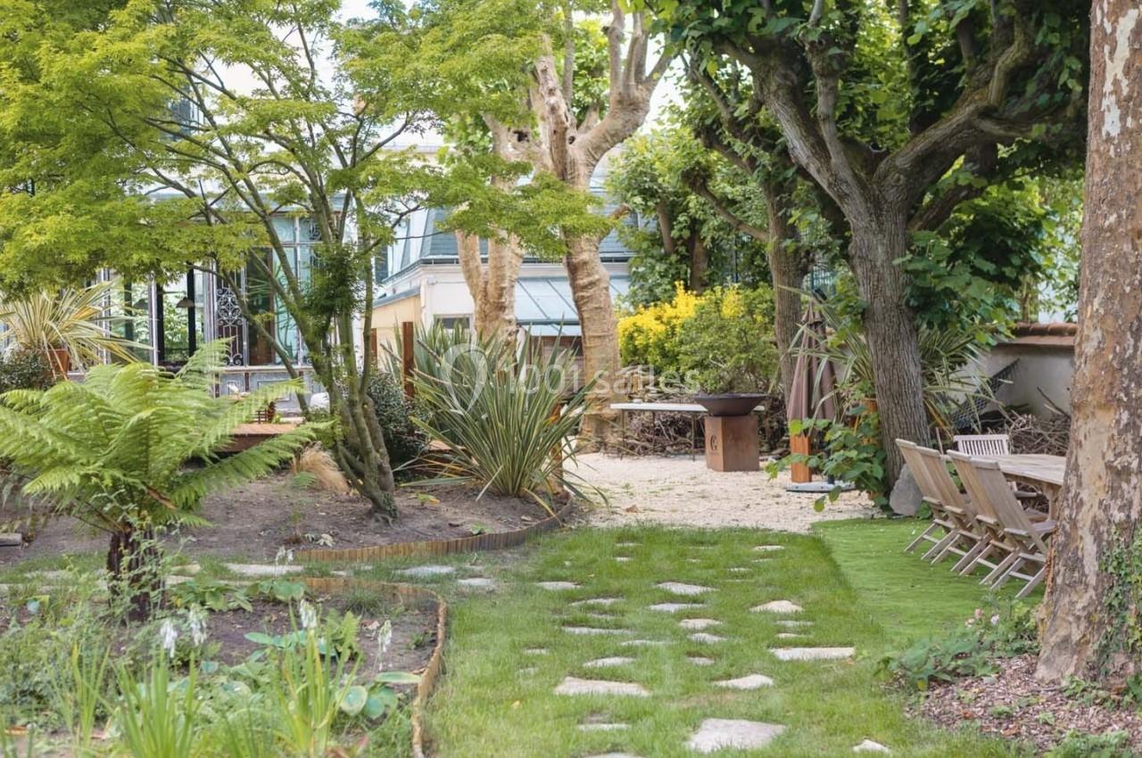 Jardin aménagé avec pelouse, allée en pierres, arbres, plantes variées, mobilier de jardin et barbecue en arrière-plan.