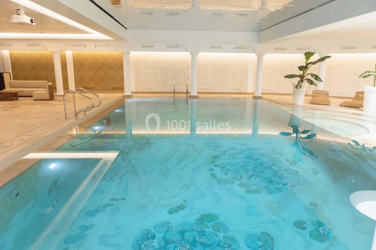 Piscine intérieure lumineuse avec eau claire, entourée de murs clairs, plantes et mobilier élégant.