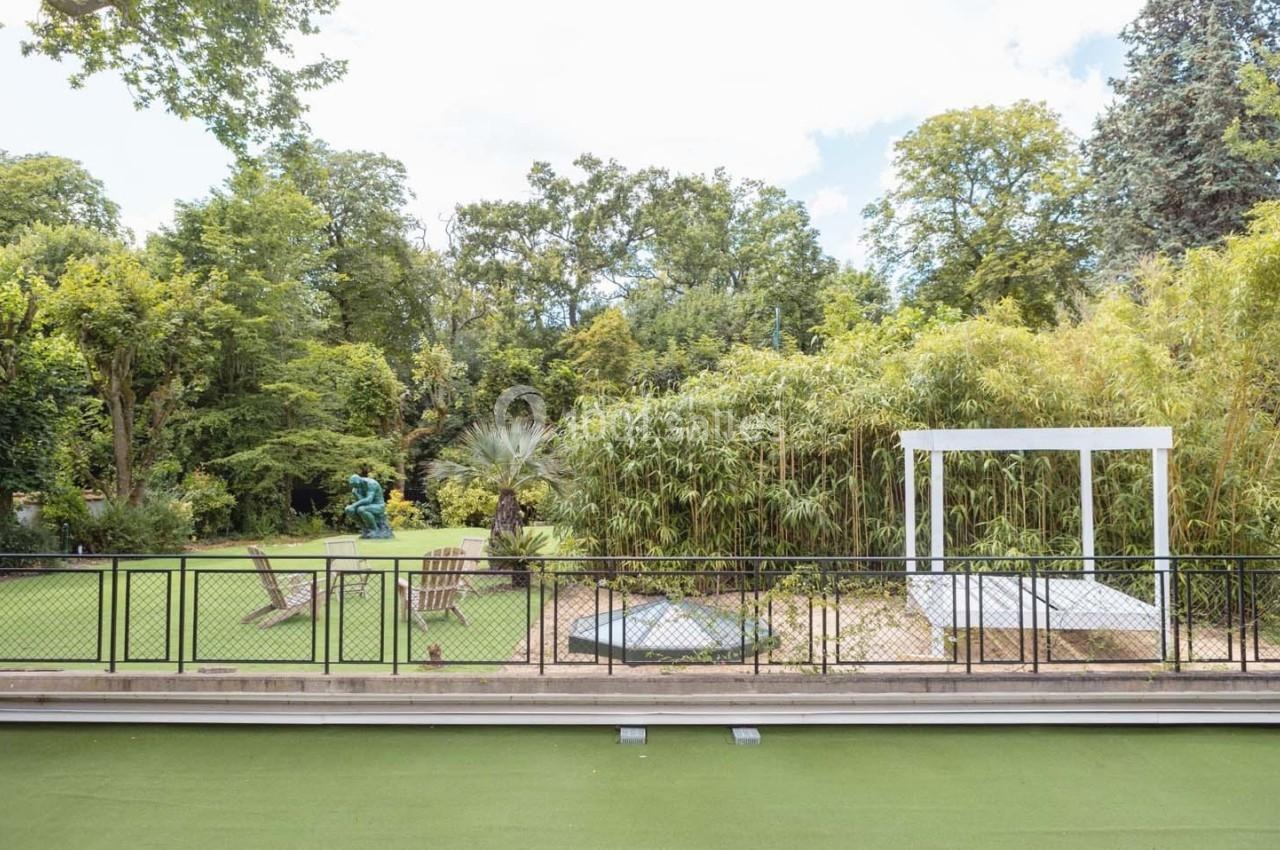 Un jardin verdoyant avec pelouse, bambous, sculptures, chaises en bois et une pergola blanche sous un ciel partiellement…