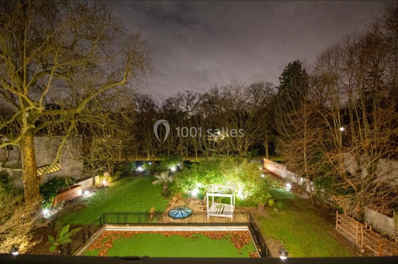 Vue nocturne d'un jardin éclairé avec pelouse, arbres dénudés, bassin central et clôtures en arrière-plan.
