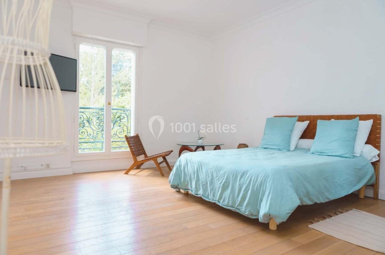 Chambre lumineuse avec lit double, parquet, fauteuil en bois, table d'appoint et grande fenêtre donnant sur un balcon.