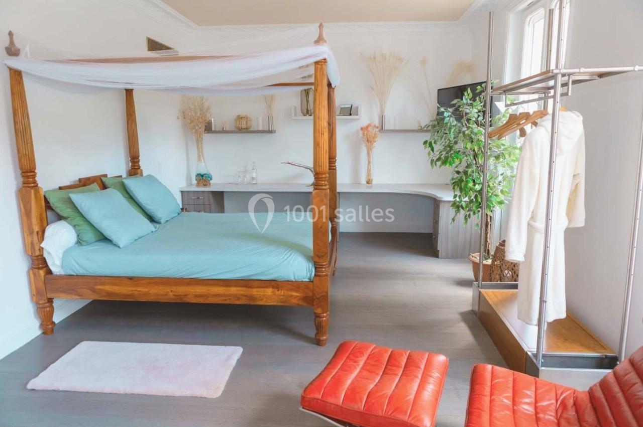 Chambre lumineuse avec lit à baldaquin en bois, bureau intégré, portant à vêtements et fauteuils en cuir rouge.