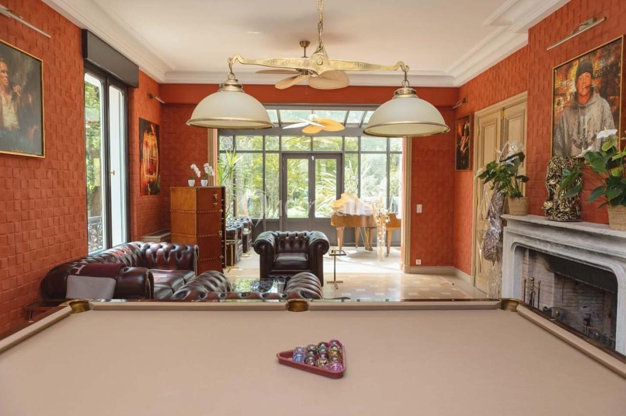 Salle avec un billard au premier plan, canapés en cuir, cheminée, et baie vitrée donnant sur un jardin.