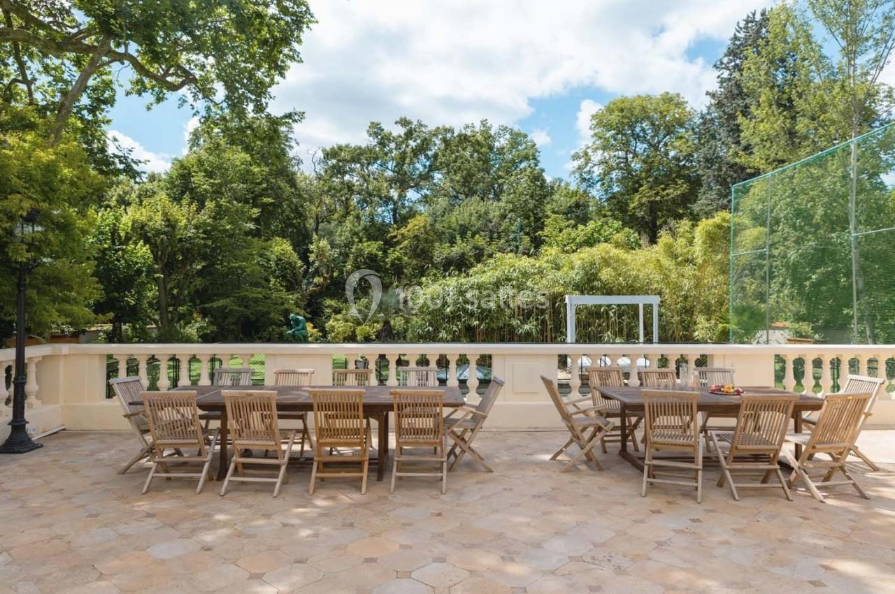 Terrasse ensoleillée avec deux grandes tables en bois entourées de chaises, donnant sur un jardin verdoyant.