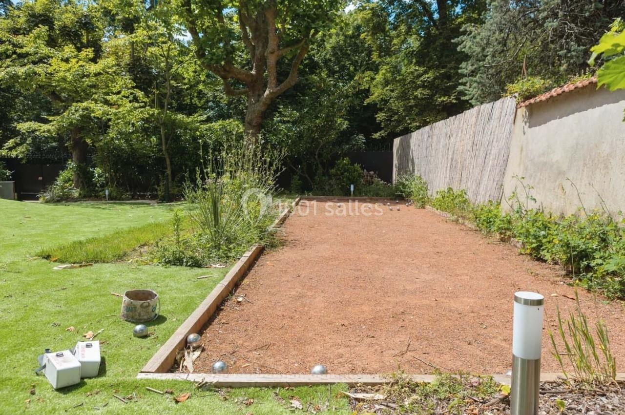 Terrain de pétanque en gravier rouge entouré de verdure et d'arbres, avec des boules dispersées au sol.