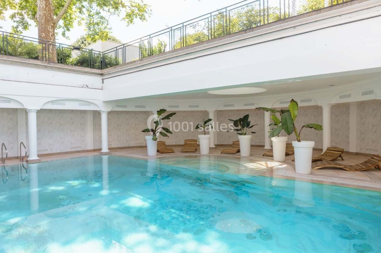 Piscine intérieure lumineuse entourée de transats en bois et de plantes en pot, avec plafond ouvert sur l'extérieur.