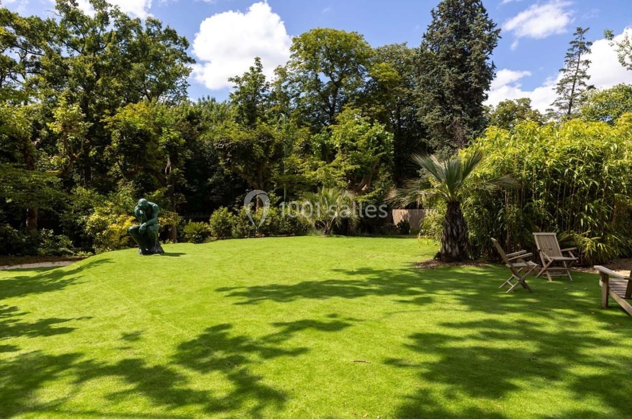 Pelouse verdoyante entourée d'arbres et de bambous, avec une statue et des chaises en bois sous un ciel dégagé.