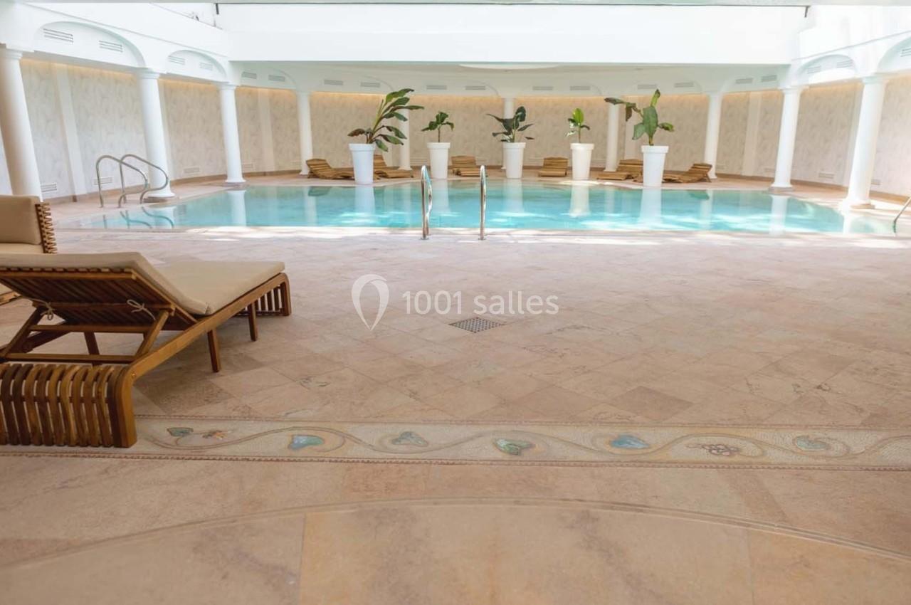 Piscine intérieure entourée de chaises longues en bois et de plantes en pots, éclairée par une lumière naturelle.