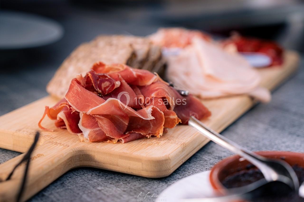 Plateau en bois avec des tranches de jambon cru, pain et autres charcuteries, posé sur une table grise.