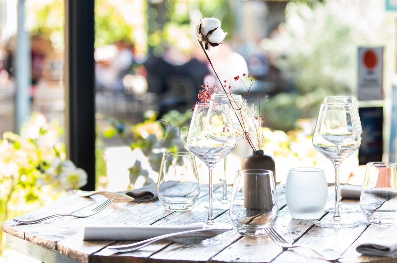 Table en bois dressée avec des verres, couverts et une décoration florale, en terrasse lumineuse.