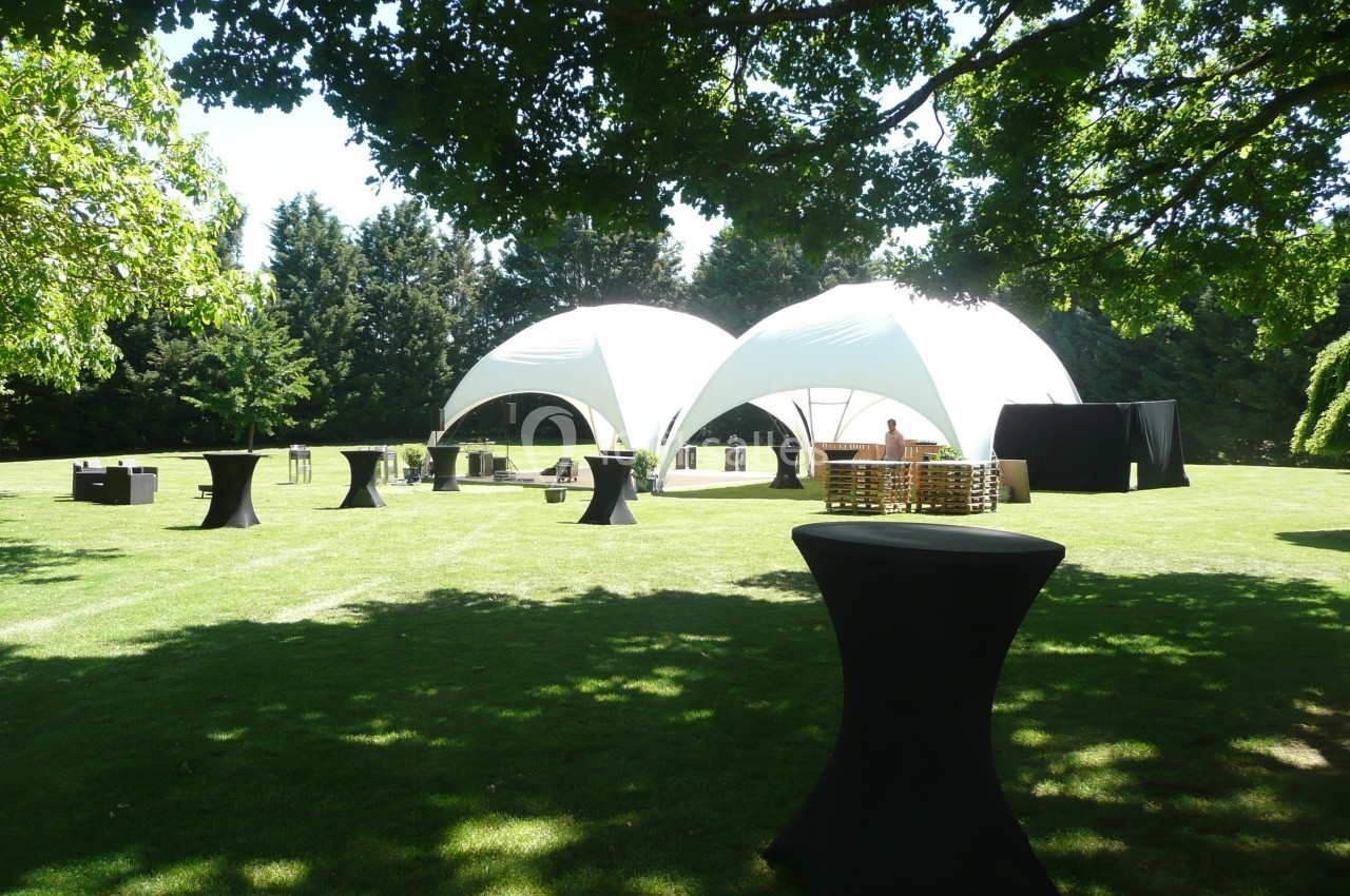 Tentes blanches modernes installées sur une pelouse avec des tables hautes noires et des arbres en arrière-plan.