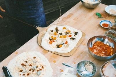 Miniature Traiteur Paris (Paris) - Pizzaiolo Tsogani #7 Un homme retire une pizza du four à l'aide d'une pelle, accompagné d'une femme souriante dans une cuisine moderne.