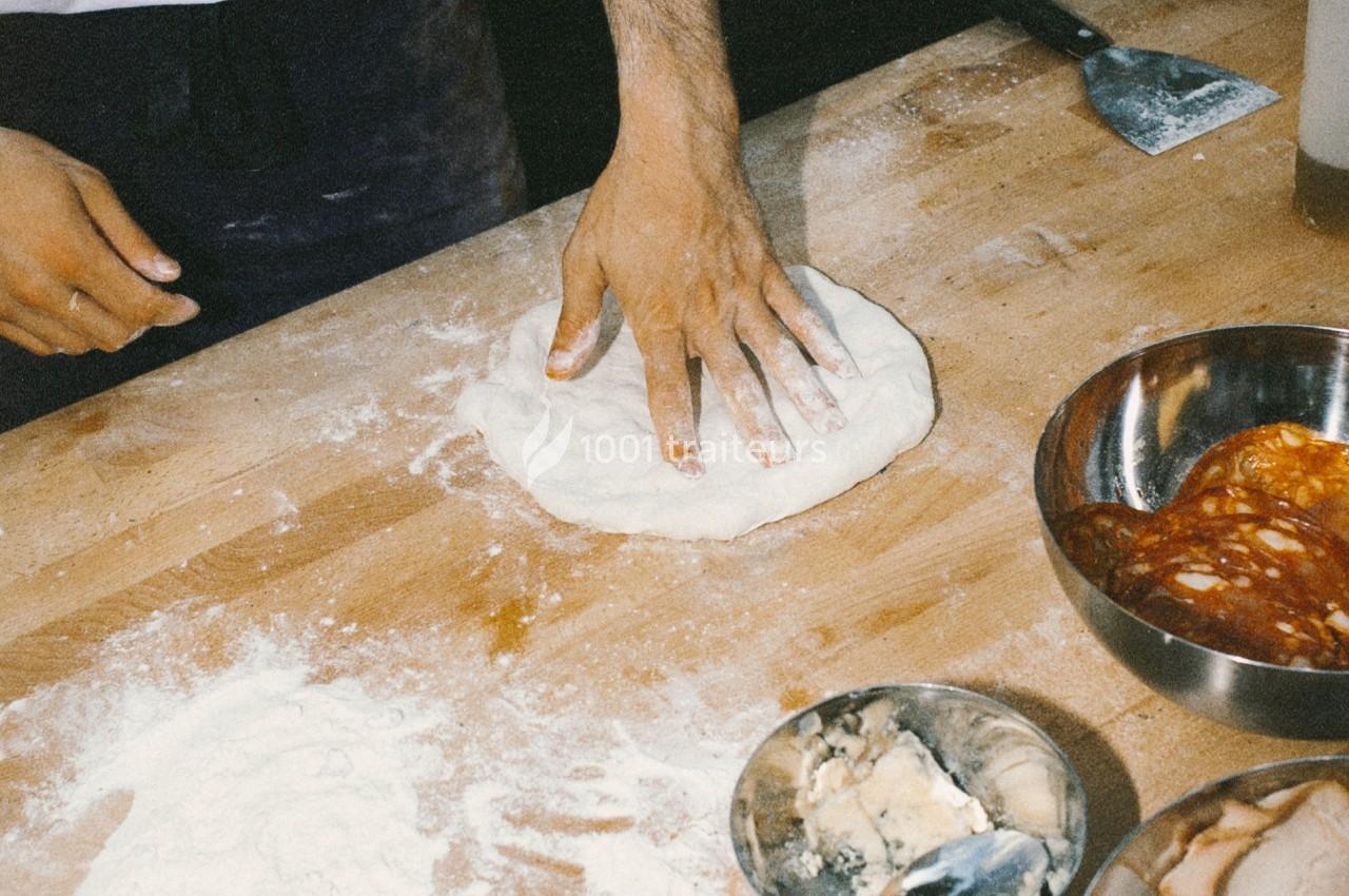 Une main aplatie une pâte à pizza sur une table en bois, entourée d'ingrédients et de farine.