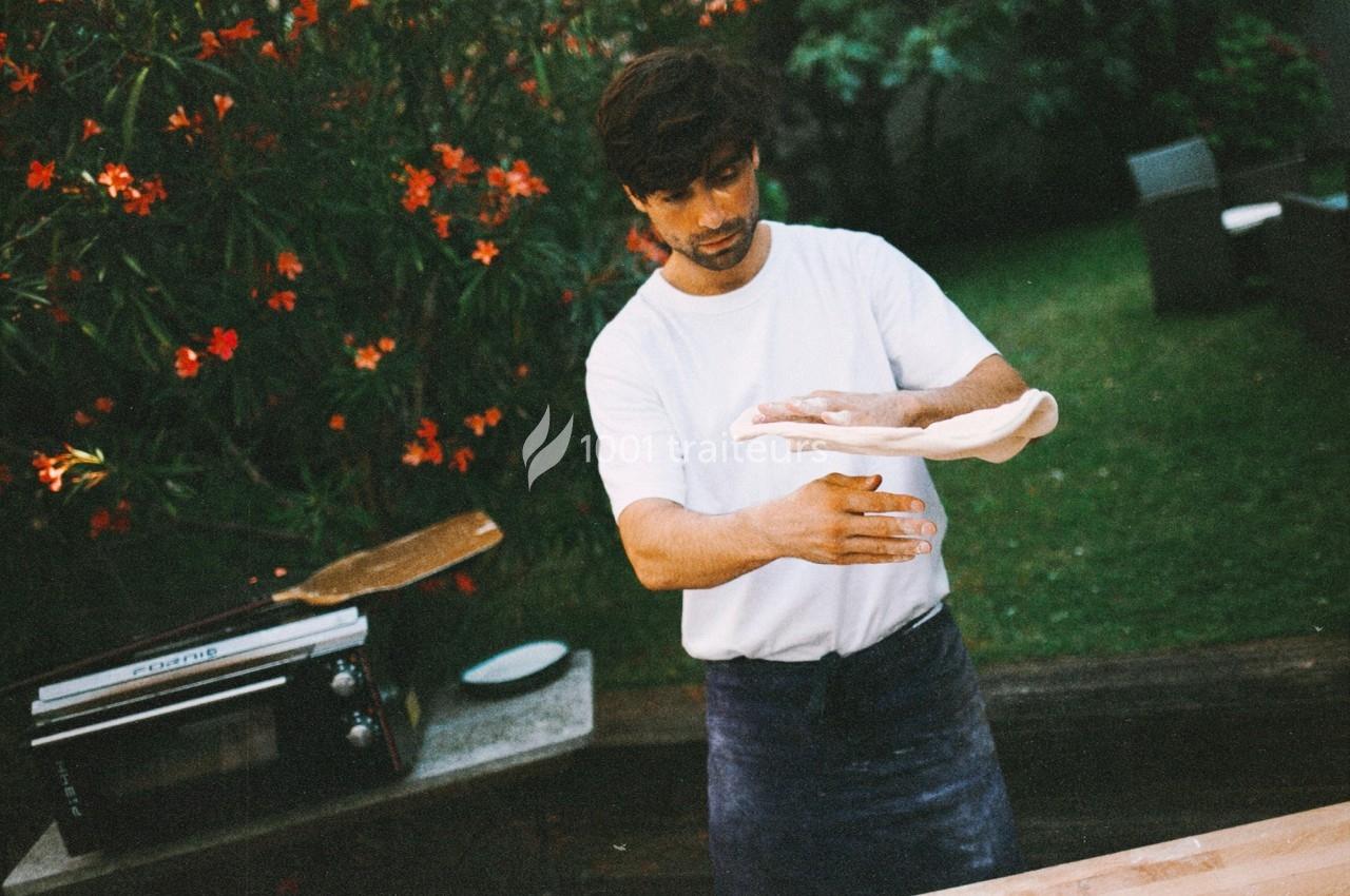 Un homme en tablier fait tourner une pâte à pizza dans un jardin avec des fleurs rouges en arrière-plan.