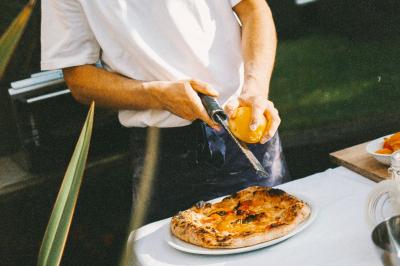 Miniature Traiteur Paris (Paris) - Pizzaiolo Tsogani #7 Un homme retire une pizza du four à l'aide d'une pelle, accompagné d'une femme souriante dans une cuisine moderne.