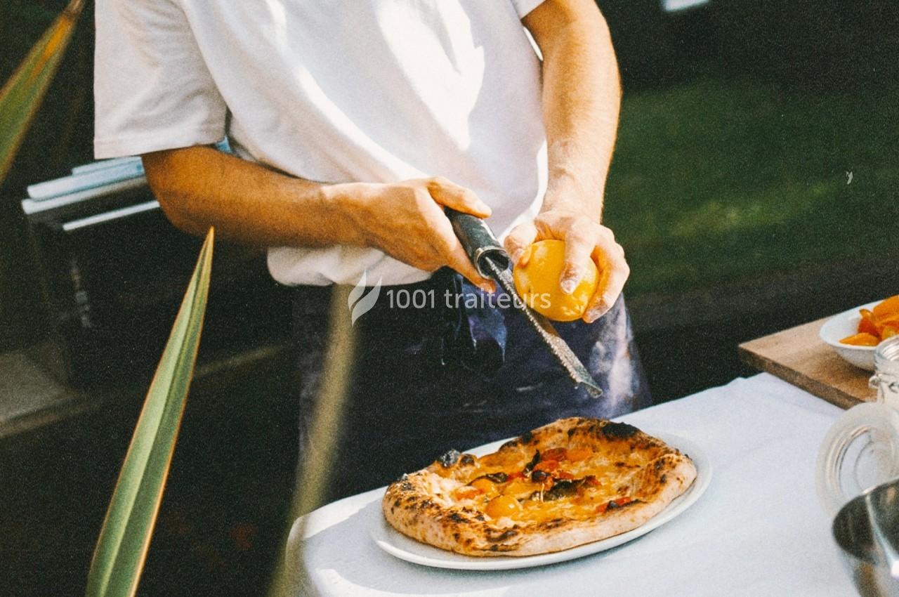Un cuisinier râpe un citron au-dessus d'une pizza garnie, posée sur une table en extérieur.
