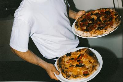 Miniature Traiteur Paris (Paris) - Pizzaiolo Tsogani #6 Un homme retire une pizza du four à l'aide d'une pelle, accompagné d'une femme souriante dans une cuisine moderne.