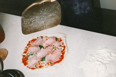 Miniature Traiteur Paris (Paris) - Pizzaiolo Tsogani #19 Un homme retire une pizza du four à l'aide d'une pelle, accompagné d'une femme souriante dans une cuisine moderne.