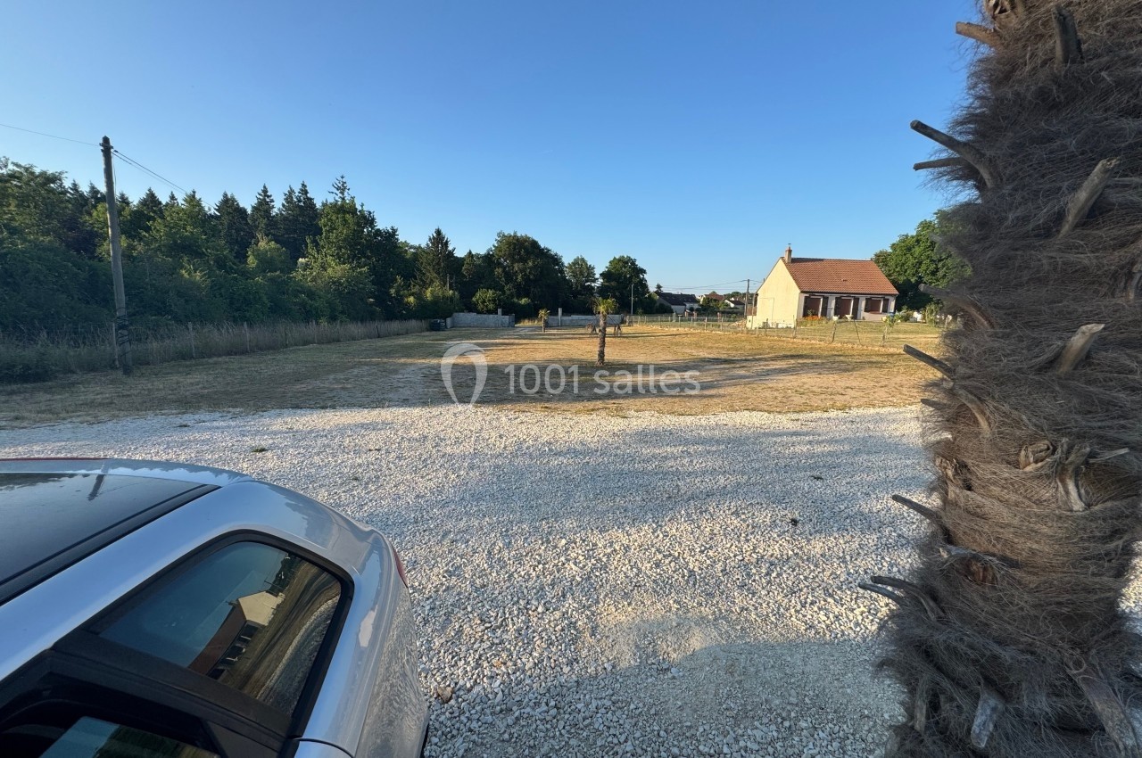 Vue d'une cour en gravier avec une voiture à gauche, un arbre au centre et une maison en arrière-plan.