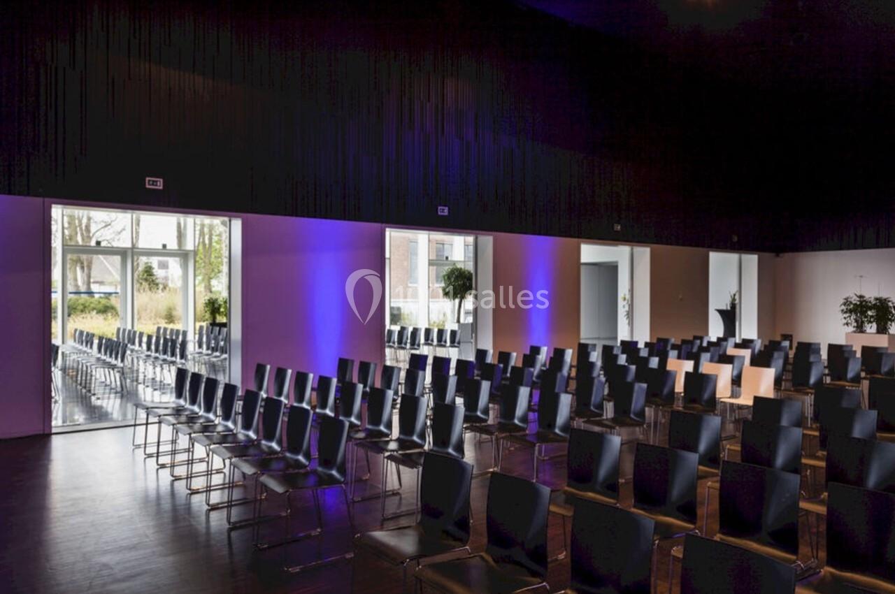 Salle de conférence moderne avec rangées de chaises noires, éclairage violet et grandes baies vitrées donnant sur l…