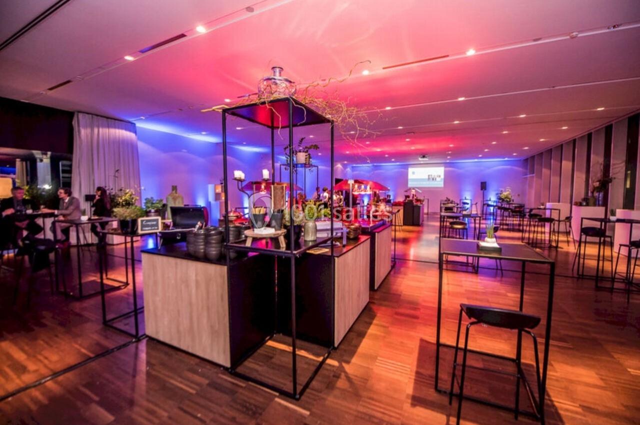 Salle de réception moderne avec éclairage coloré, tables hautes et buffet central décoré de plantes.