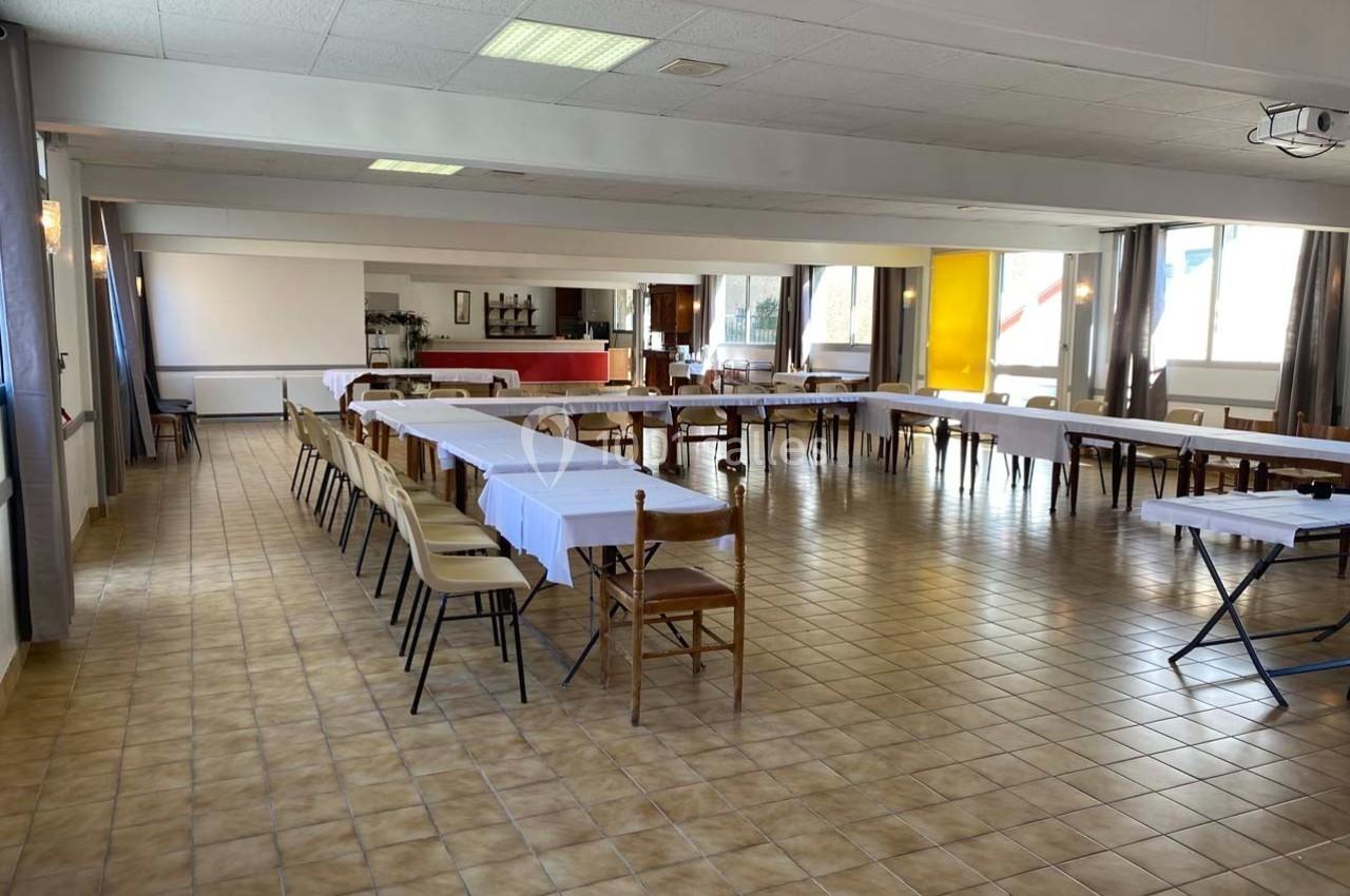 Salle spacieuse avec tables rectangulaires alignées et chaises, éclairée par des fenêtres et des lumières au plafond.