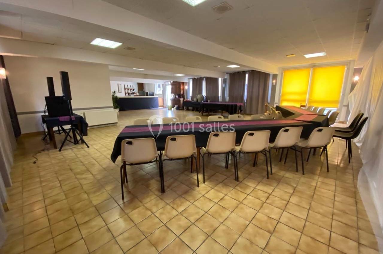 Salle de réunion avec tables disposées en U, chaises alignées, sol carrelé et grandes fenêtres avec stores jaunes.
